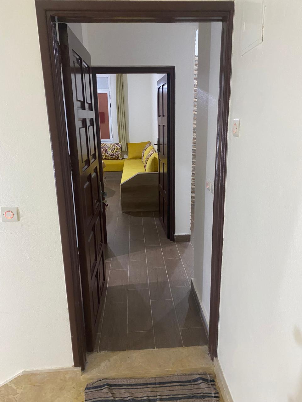 A hallway leads into a spacious living area, with double doors visible on each side and a glimpse of a yellow sofa in the background. The walls are painted in neutral colors, and the floor features a mix of tiles, enhancing the overall layout.
