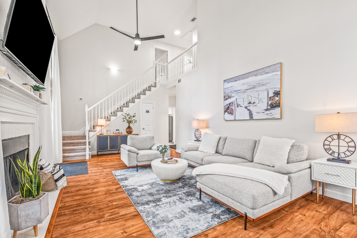 A spacious living area features a light-colored sectional sofa positioned around a circular coffee table. An area rug complements the wood flooring, while natural light floods the room from large windows. A fireplace and minimalist decor add to the inviting ambiance.