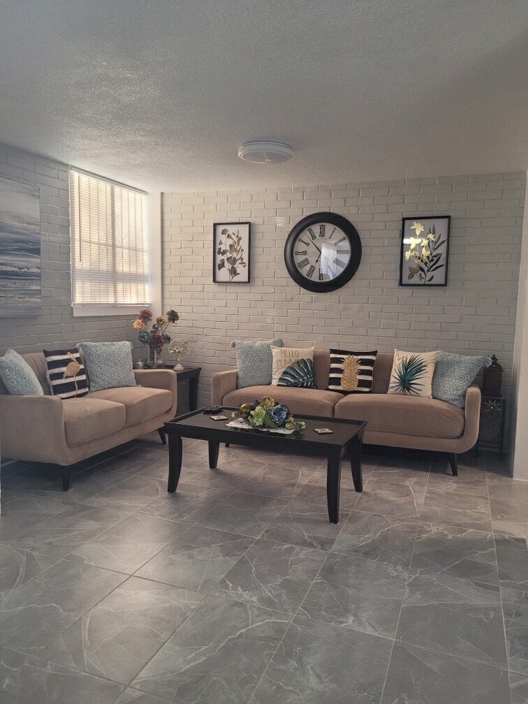 A cozy living area features a light-colored sofa adorned with assorted cushions and a decorative table. Wall art with botanical themes complements the space, while a large clock adds functionality. Natural light filters through a window, highlighting the modern tile flooring.