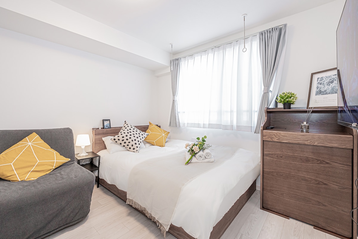 A bright and modern room is illuminated by natural light from a large window. Two spacious beds with neutral bedding are complemented by decorative pillows in vibrant colors. A cozy sofa and a dresser add functionality to the space, while a small plant brings a touch of nature.