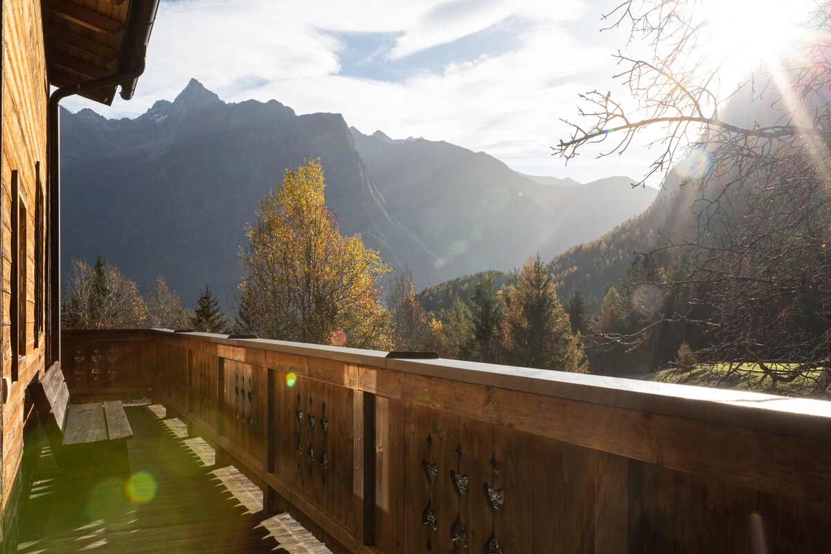 A wooden balcony offers a scenic view of majestic mountains and lush greenery, illuminated by the soft glow of sunlight. The space features a simple wooden bench, inviting relaxation while taking in the surrounding natural beauty.