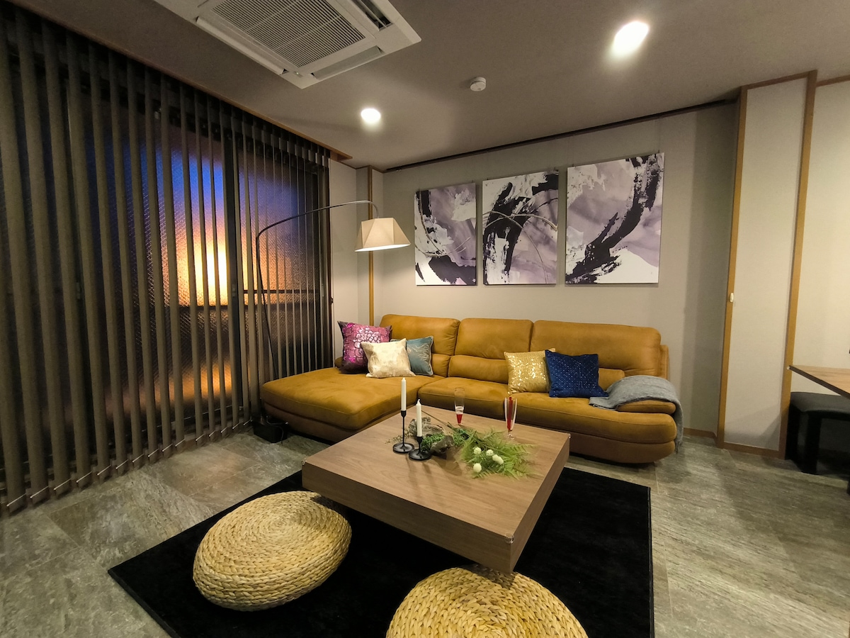 A modern living space features a tan sectional sofa adorned with decorative cushions in various colors. A low coffee table sits on a dark rug, complemented by minimal decor. Natural light filters through the sheer curtains, enhancing the contemporary ambiance.