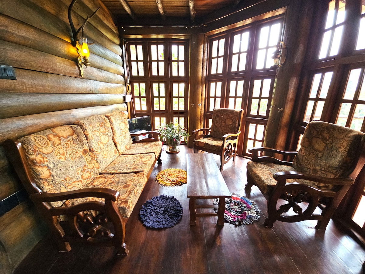 A cozy sitting area features two upholstered armchairs and a wooden couch with intricate carvings. A wooden coffee table is centered on a polished floor, surrounded by colorful round rugs. Large windows allow ample natural light and provide views of the lush surroundings.