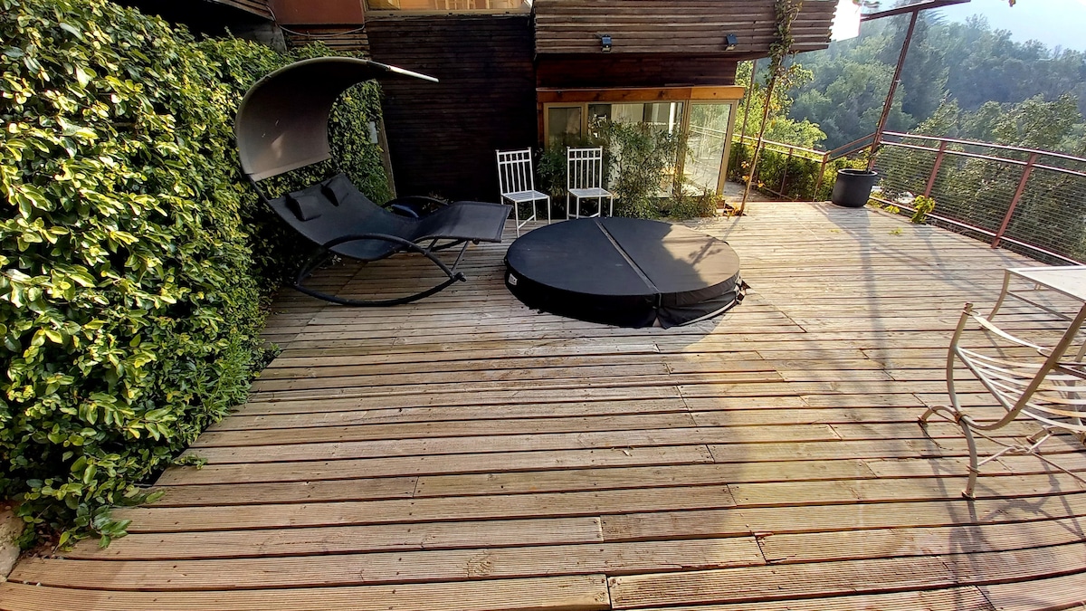 A spacious wooden deck is presented, featuring a circular black cover and a lounge chair with a unique design. Two white chairs are positioned nearby, and lush greenery surrounds the area, enhancing the outdoor setting.