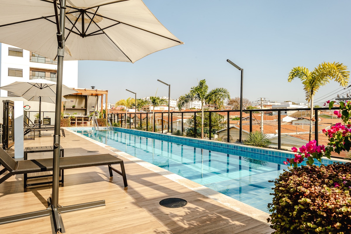 The rooftop pool area features a long pool bordered by lounge chairs and large umbrellas for shade. Surrounding greenery and vibrant flowers enhance the outdoor ambiance. The view includes a skyline of buildings and clear blue sky, creating a relaxing environment.