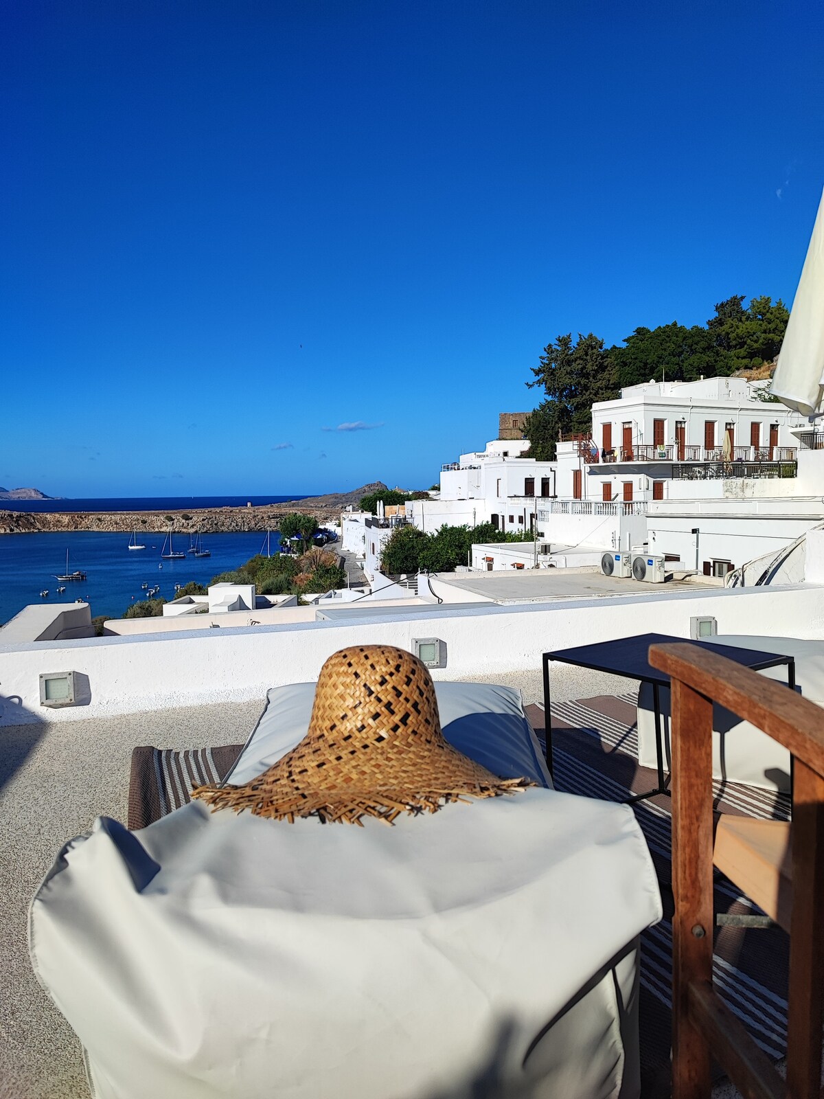 A sunlit terrace offers views of the sea and surrounding white buildings. A stylish sunbed is positioned for relaxation, with a woven sunhat resting on it. A small table stands nearby, completing the serene outdoor space against a backdrop of clear blue skies.