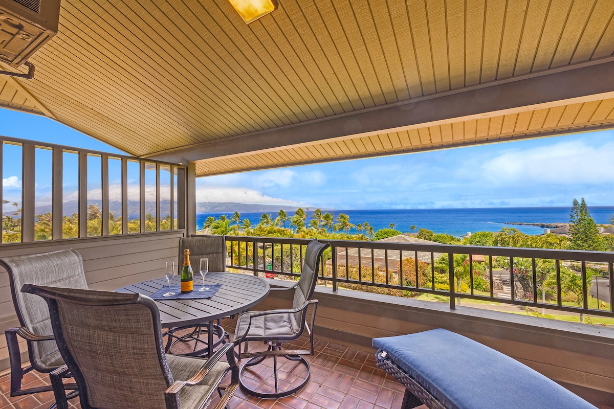 An outdoor lanai features a dining table with four chairs and a lounge chair, overlooking expansive ocean views. Lush greenery can be seen in the foreground, while distant islands are visible on the horizon beneath a clear blue sky.