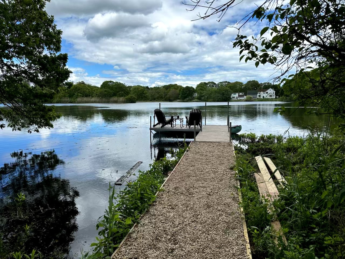 A gravel pathway leads to a wooden dock extending over the serene lake. Two chairs are positioned on the dock, overlooking calm waters that reflect the surrounding greenery and cloudy sky. Lush foliage frames the scene, enhancing the natural beauty.