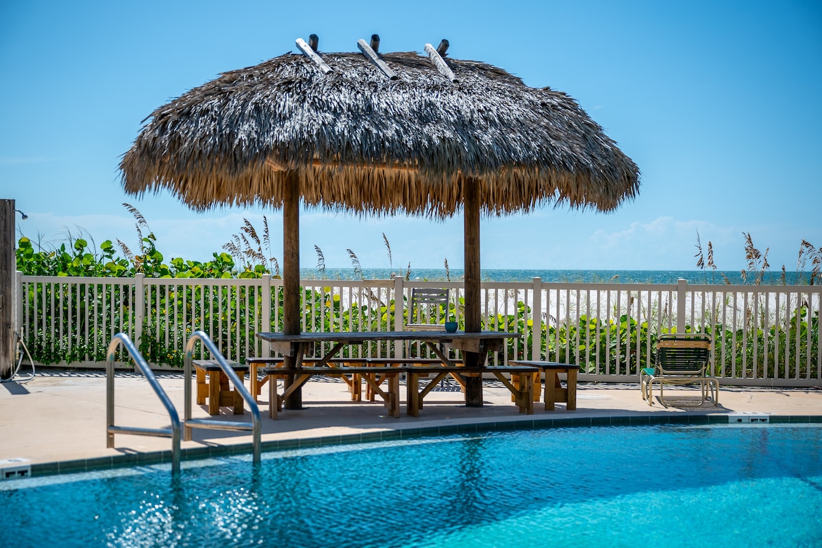 A thatched-roof pavilion with a picnic table is set by the poolside, offering a shaded spot for gatherings. The shimmering pool reflects clear blue skies, and the beach is visible in the background, framed by lush greenery and swaying grasses.