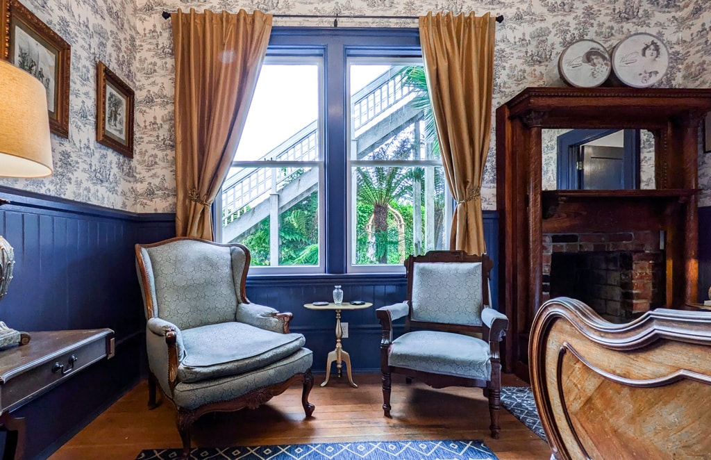 A seating area is furnished with two upholstered chairs and a small table, placed beside a large window dressed with elegant drapery. The room features patterned wallpaper and wooden accents, creating a cozy and historical ambiance.