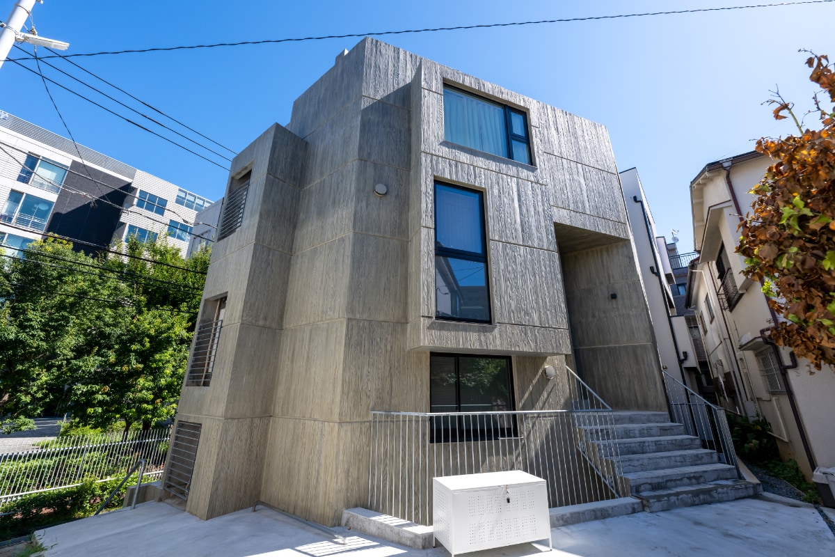 The modern exterior of the building features a textured concrete façade, characterized by angular lines and large windows. A set of concrete steps leads to the entrance, while the surrounding area includes greenery and neighboring residential structures.