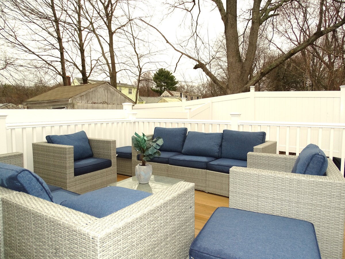 A private outdoor deck features a seating area furnished with a woven sectional sofa and matching armchairs, all adorned with blue cushions. A coffee table sits in the center, complemented by a decorative plant. Surrounding trees and a white fence provide privacy.