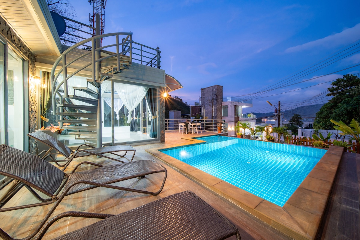 An outdoor area is presented featuring a private pool with clear blue water. A spiral staircase leads to a second level, while loungers are positioned nearby. The space is illuminated softly by ambient lighting as dusk approaches, enhancing the inviting atmosphere of the villa.