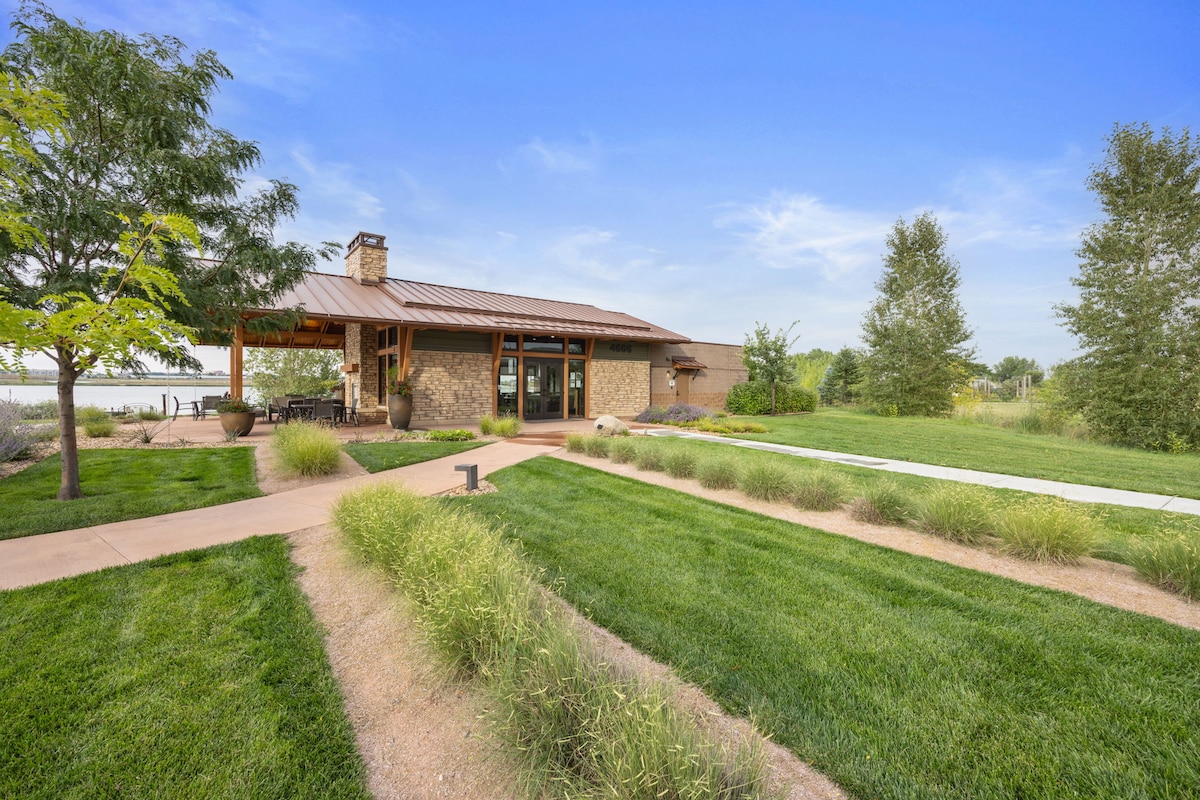 A modern clubhouse features a stone exterior and large windows, surrounded by landscaped gardens. A pathway of well-maintained grass leads to the entrance, offering a sense of openness and connection to nature, with trees and shrubs providing additional greenery.