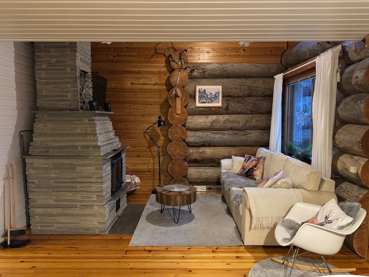 A spacious living area features a comfortable sofa arranged around a circular coffee table. A stone chimney rises beside the seating, complementing the wooden walls. Natural light filters through a large window, partially covered by light curtains. Decorative wall art adds character to the space.