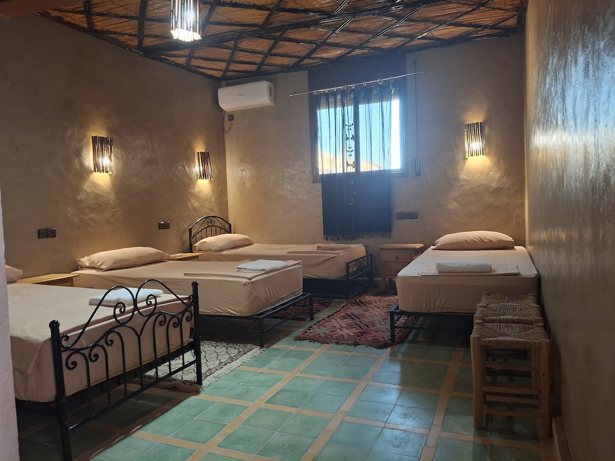 A spacious guest room features multiple beds, arranged neatly with linens and towels. Soft lighting is provided by wall sconces, and a window allows natural light to fill the space. Textured walls and traditional decor contribute to a warm and inviting atmosphere.