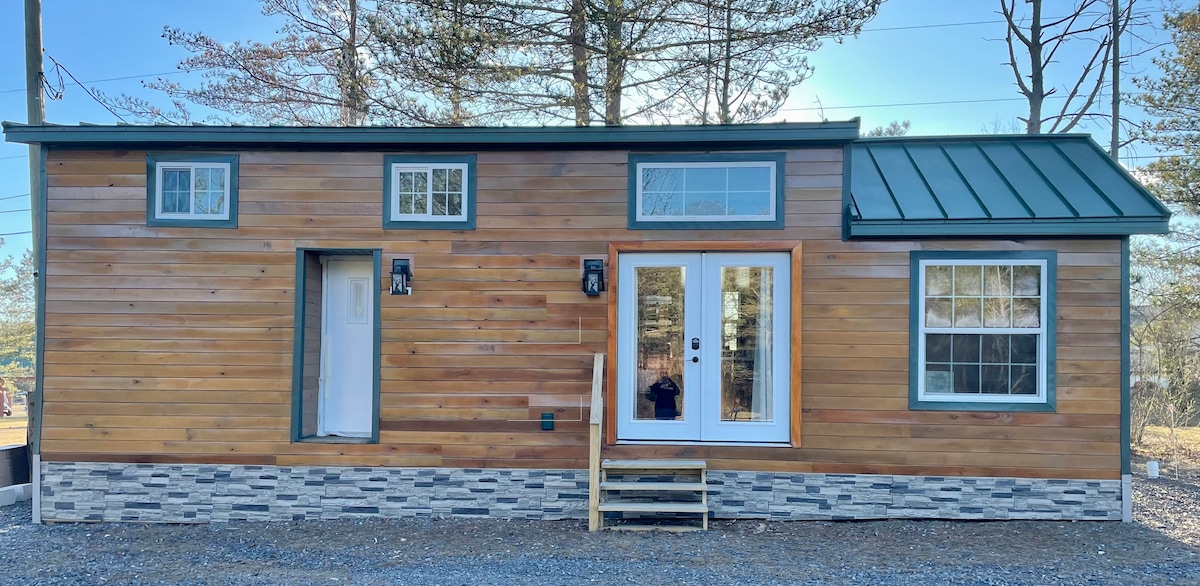 The exterior of a cabin-style tiny home is shown, featuring wood paneling and stone detailing. Large windows allow natural light to enter, and a small set of steps leads to double front doors. The surrounding area includes trees and gravel.