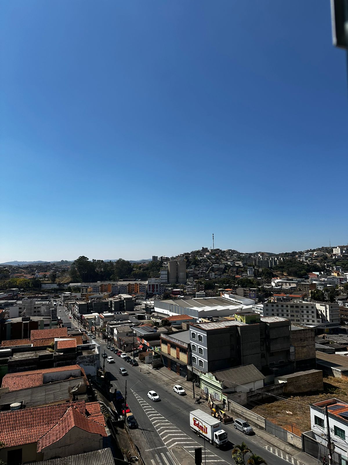 A panoramic urban view showcases a clear blue sky above a vibrant city landscape. Numerous buildings and rooftops are visible, along with streets and vehicles below. In the background, a distant hillside is seen, featuring structures that rise against the horizon.