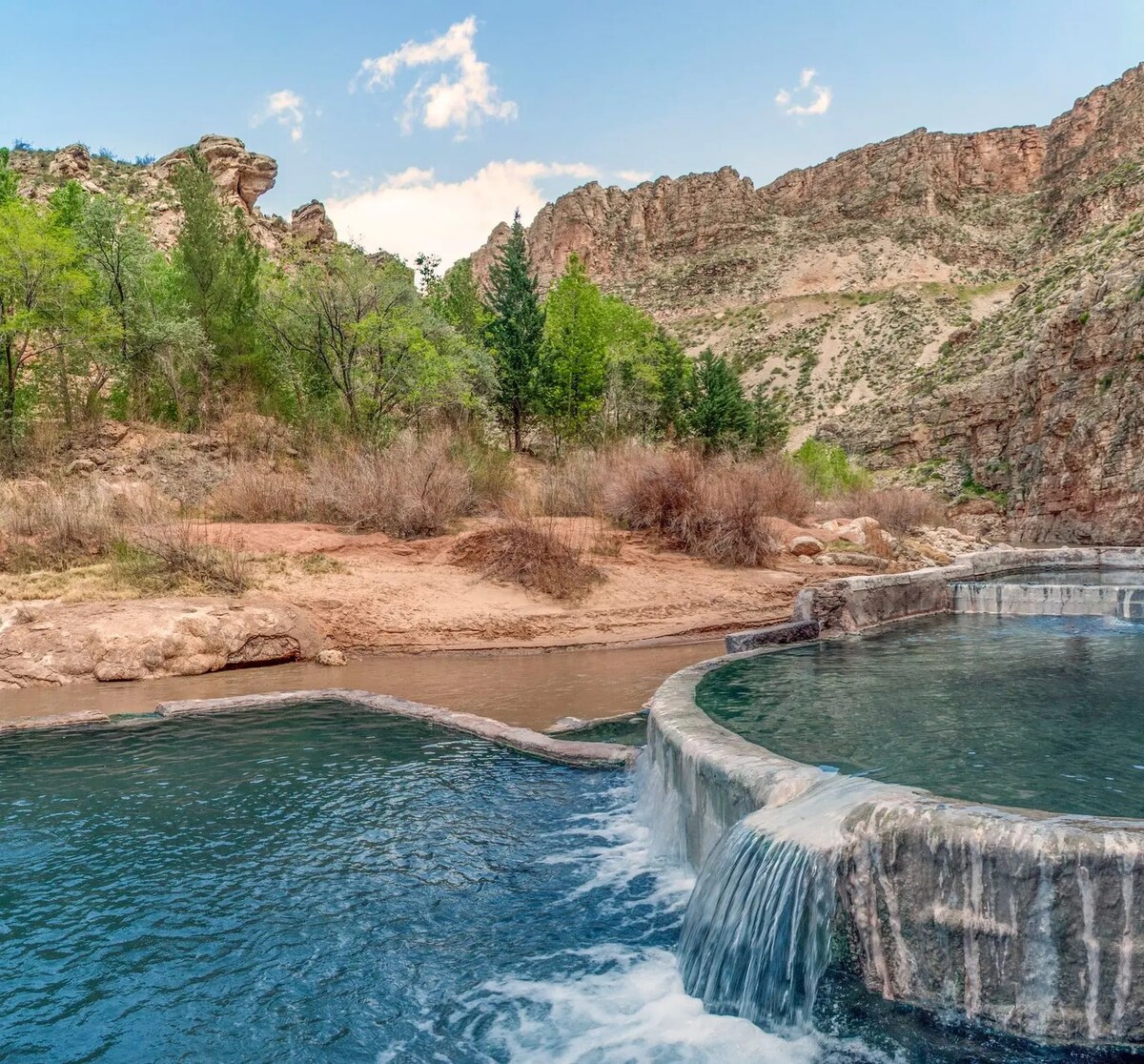 A scenic view of a natural hot springs pool is complemented by rocky hills and lush greenery in the background. The gently flowing water cascades over a stone edge, creating a serene atmosphere, with rugged terrain and distant cliffs framing the setting.