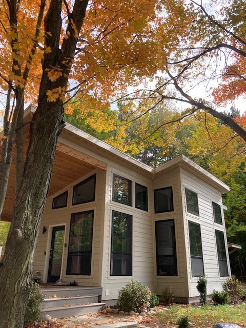 The modern cottage features large windows framed by autumn-colored foliage, allowing natural light to highlight its architecture. A welcoming entrance is complemented by steps leading up to a covered porch, creating an inviting outdoor space amidst the serene wooded setting.