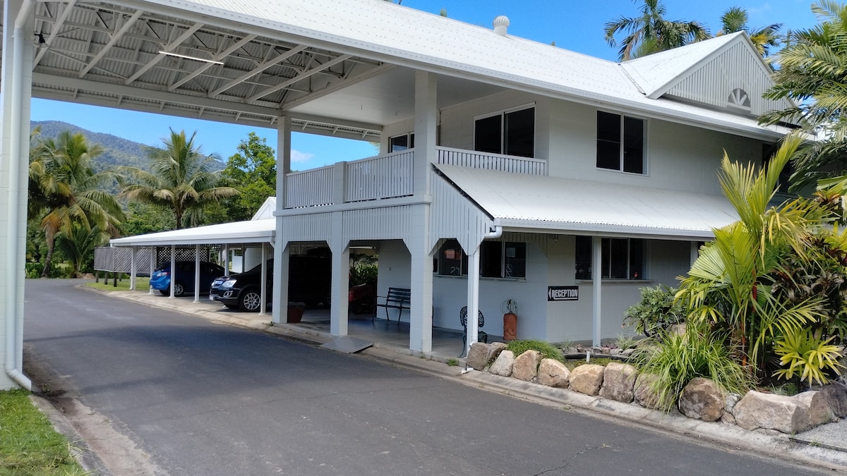 A two-story building with a covered entrance is shown, surrounded by tropical gardens. Multiple parking spaces are visible, along with vehicles parked underneath the structure. Lush greenery and palm trees frame the scene, providing a serene environment.