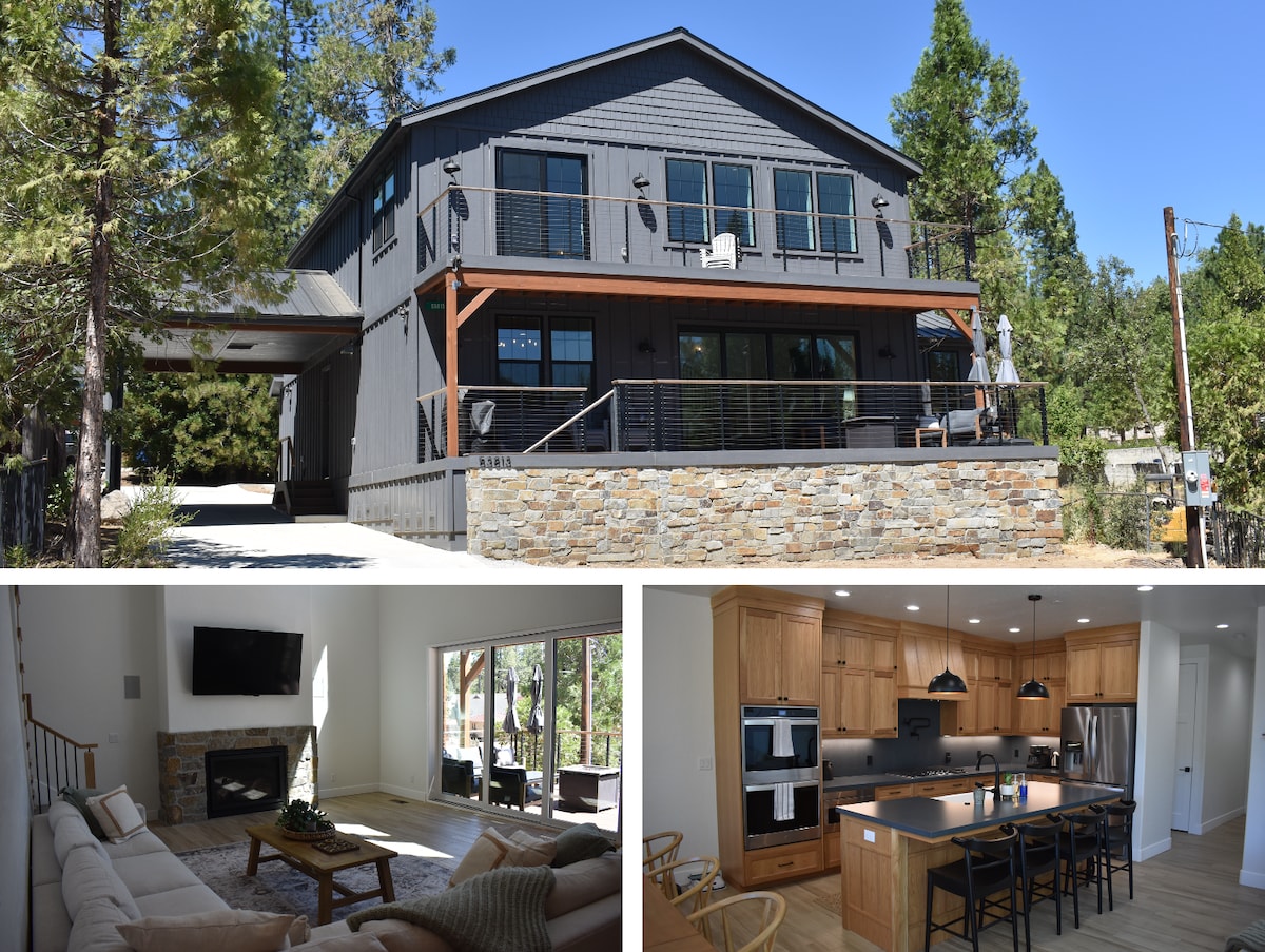 The exterior of a contemporary home is shown, featuring a dark wood and stone facade. A spacious porch extends across the front, complemented by large windows. Nearby trees offer natural shade, and a driveway leads up to the entrance.