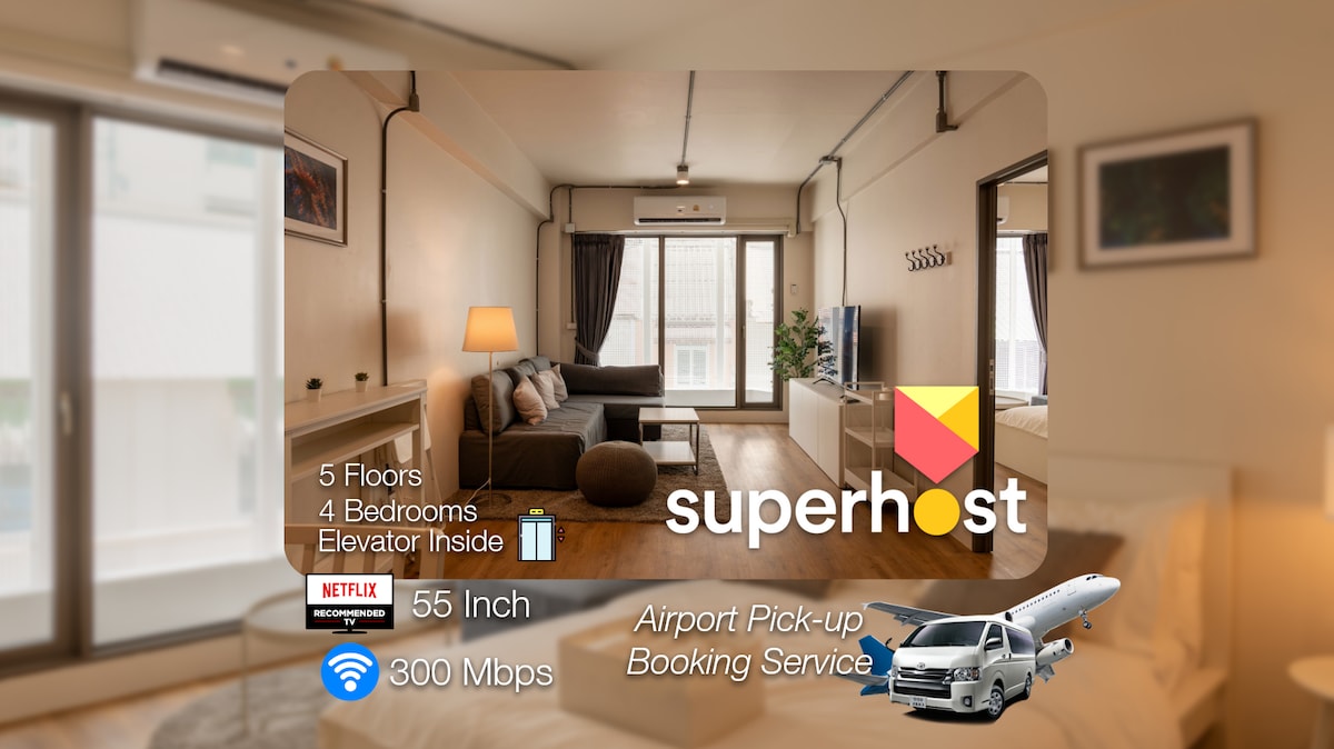 A spacious living area is furnished with a comfortable sofa and a 55-inch TV mounted on the wall. Natural light floods through large windows, offering views of the surrounding neighborhood. The room features an organized layout with light-colored decor and wooden flooring.