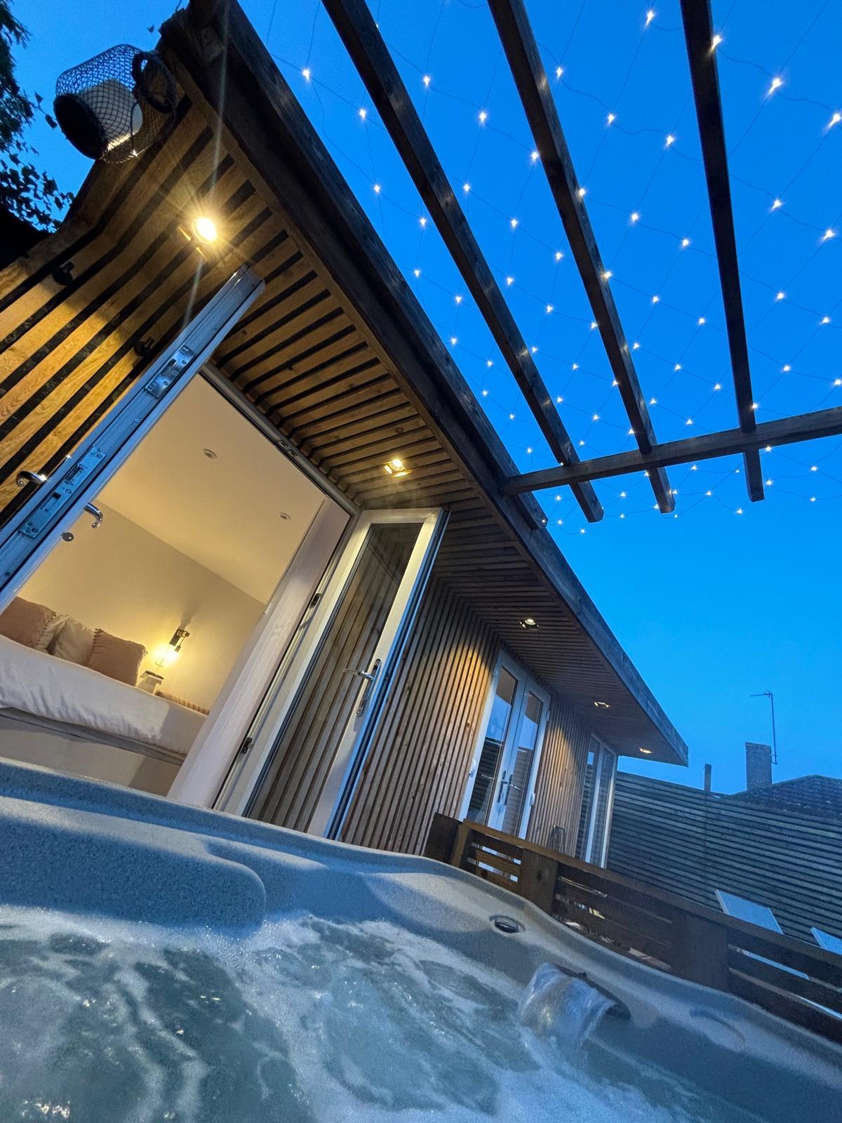 A private hot tub is observed in the foreground, with gentle bubbling water. In the background, a modern log cabin structure is visible, illuminated by warm outdoor lighting. Above, a starry sky is complemented by string lights, creating a serene evening ambiance.