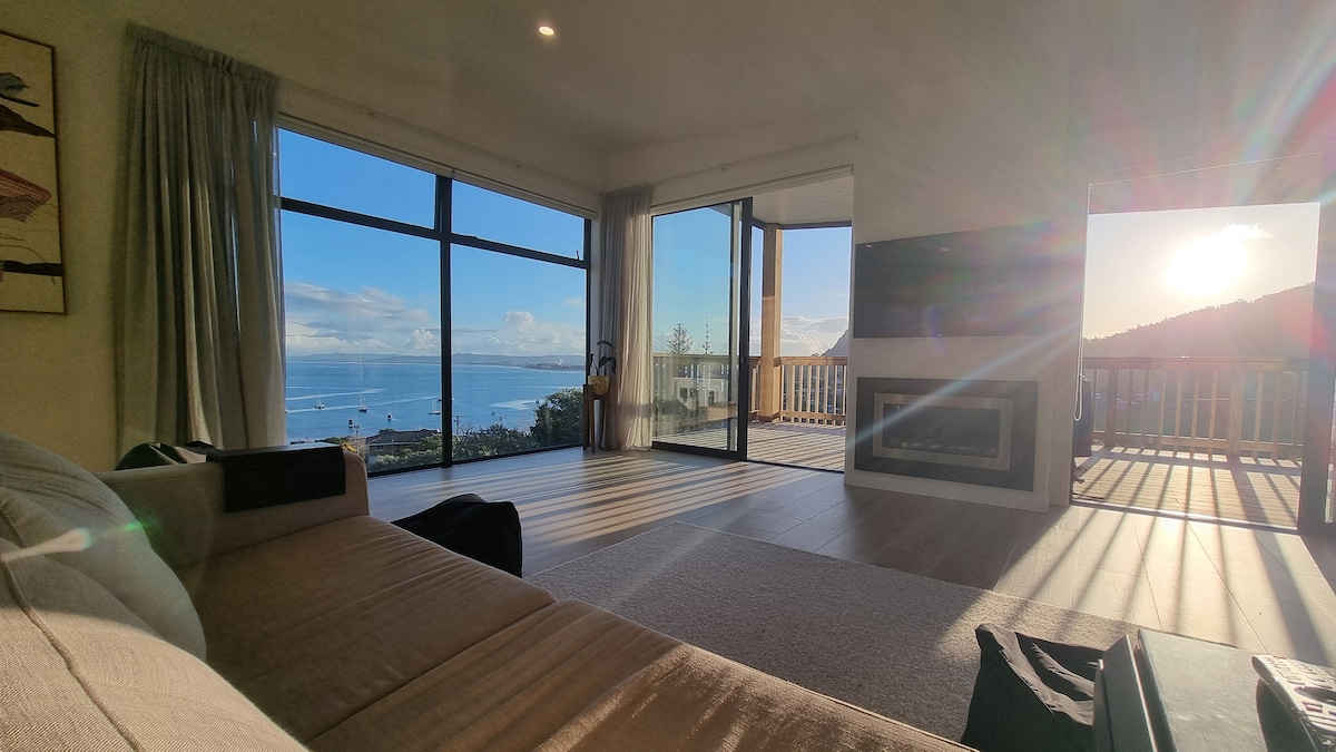 A lounge area is presented with large glass doors offering expansive views of Bream Bay and Mount Manaia. Natural light fills the space, reflecting off the walls and flooring. A comfortable sofa and a gas fireplace enhance the inviting atmosphere.