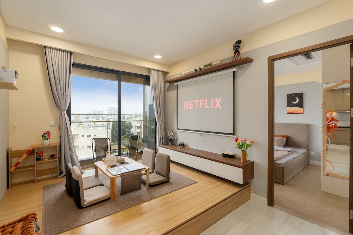 A modern living area features a large window allowing natural light to fill the space. A wall-mounted screen displays the Netflix logo. A low table is surrounded by four chairs, and a comfortable seating area is positioned nearby, enhancing the welcoming ambiance.