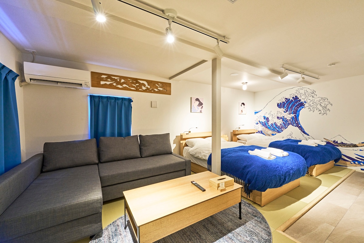 A spacious room is presented with two double beds adorned in blue covers, complemented by a sofa in a muted gray color. A wooden coffee table rests in the center, while artworks inspired by traditional Japanese themes are visible on the walls.