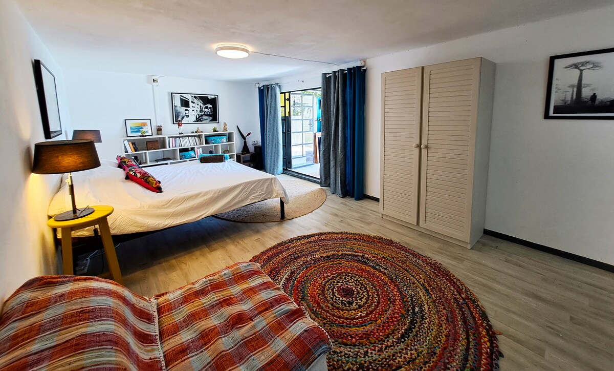 A spacious studio room features a large bed covered with a light duvet, accompanied by a colorful woven rug. A cozy seating area with a striped blanket is set beside a sliding glass door that opens to the garden. A light-colored wardrobe and artwork adorn the walls.