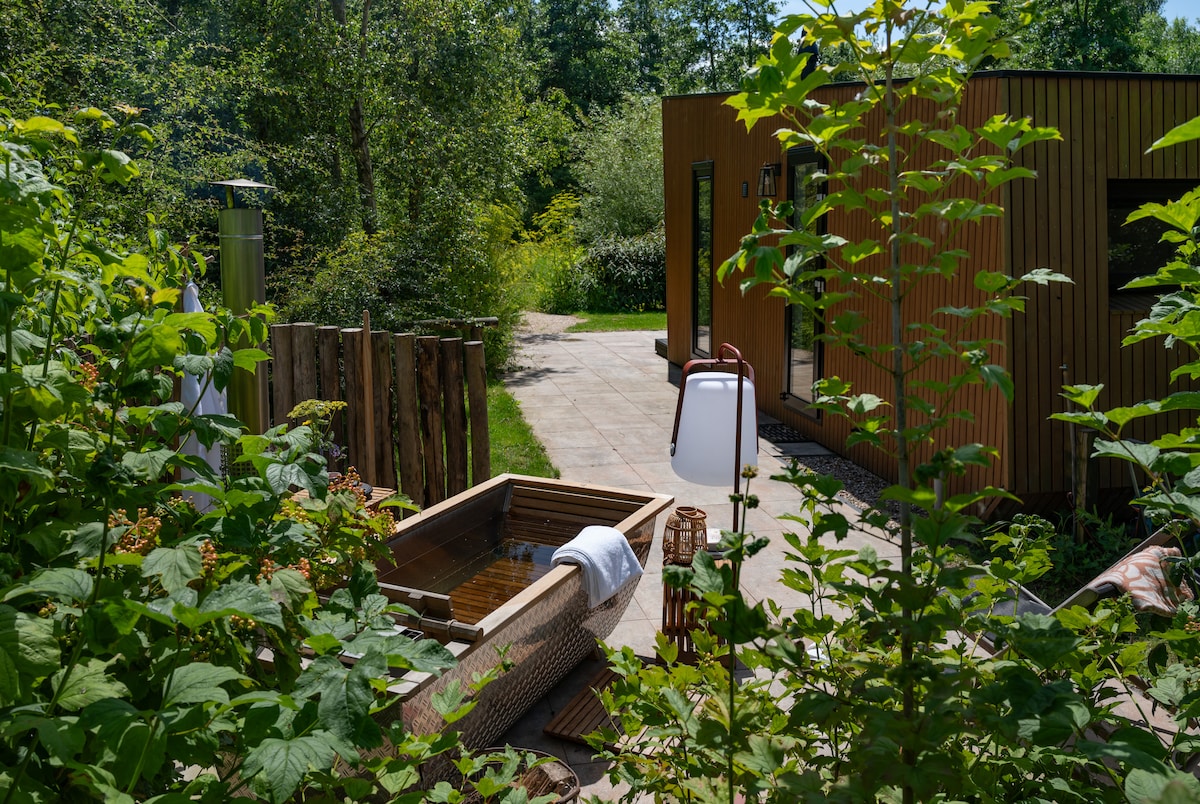 A private wooden hot tub is surrounded by lush greenery, with a clear pathway leading to a lodge structure. The soothing water invites relaxation, while trees frame the serene outdoor space, enhancing the lodge's natural setting.