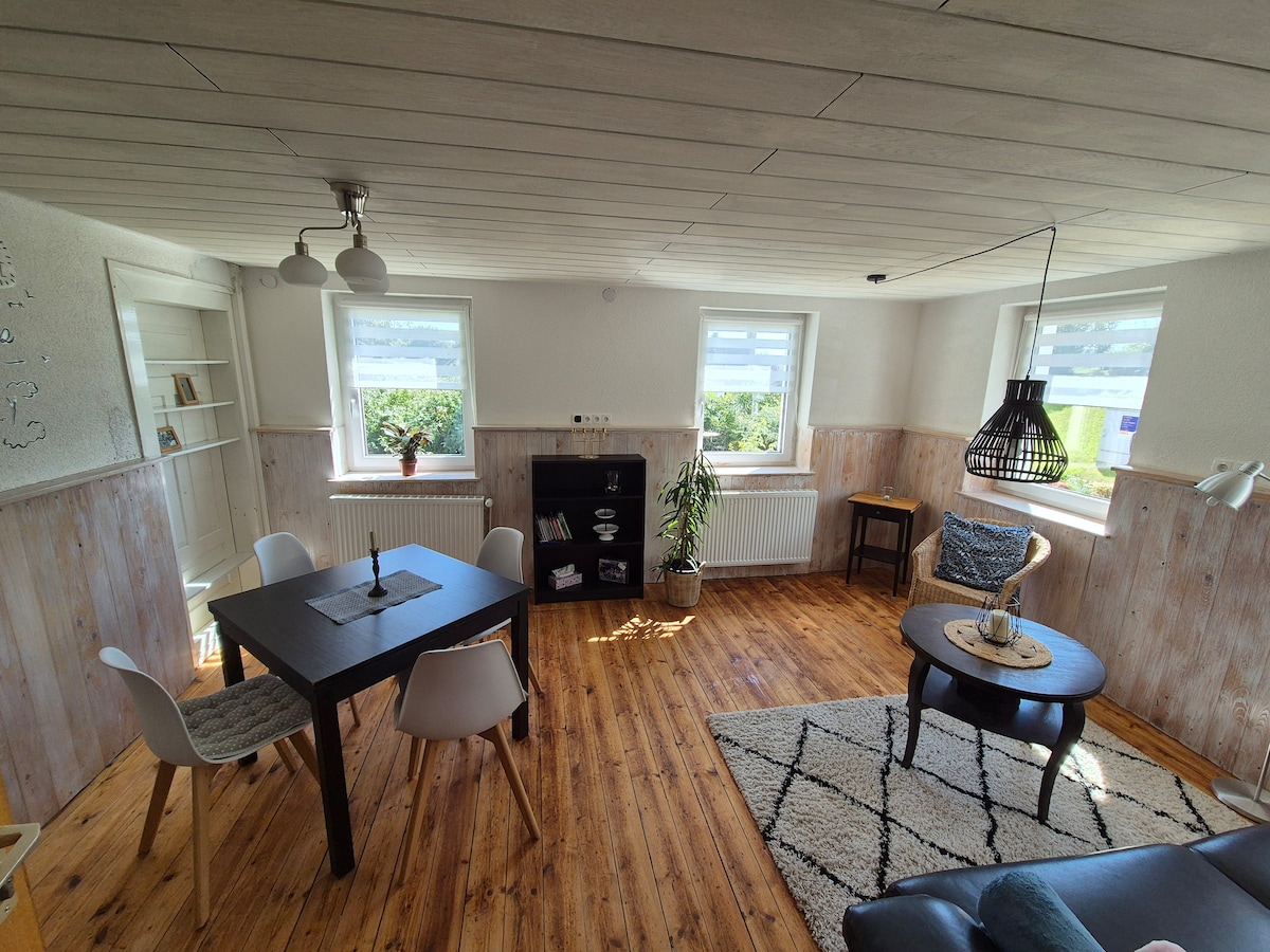 A bright and inviting living area features wooden flooring and walls. Two windows allow natural light to fill the space. A black dining table is surrounded by four chairs, while a cozy sitting area includes a dark couch and a side table. A decorative plant adds a touch of greenery.