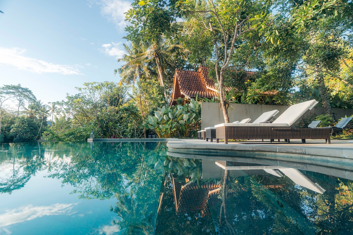 An infinity pool is surrounded by lush tropical greenery, reflecting the clear blue sky above. Comfortable loungers are positioned beside the water, inviting relaxation. In the distance, a traditional Balinese structure with a terracotta roof is nestled among the vibrant foliage.
