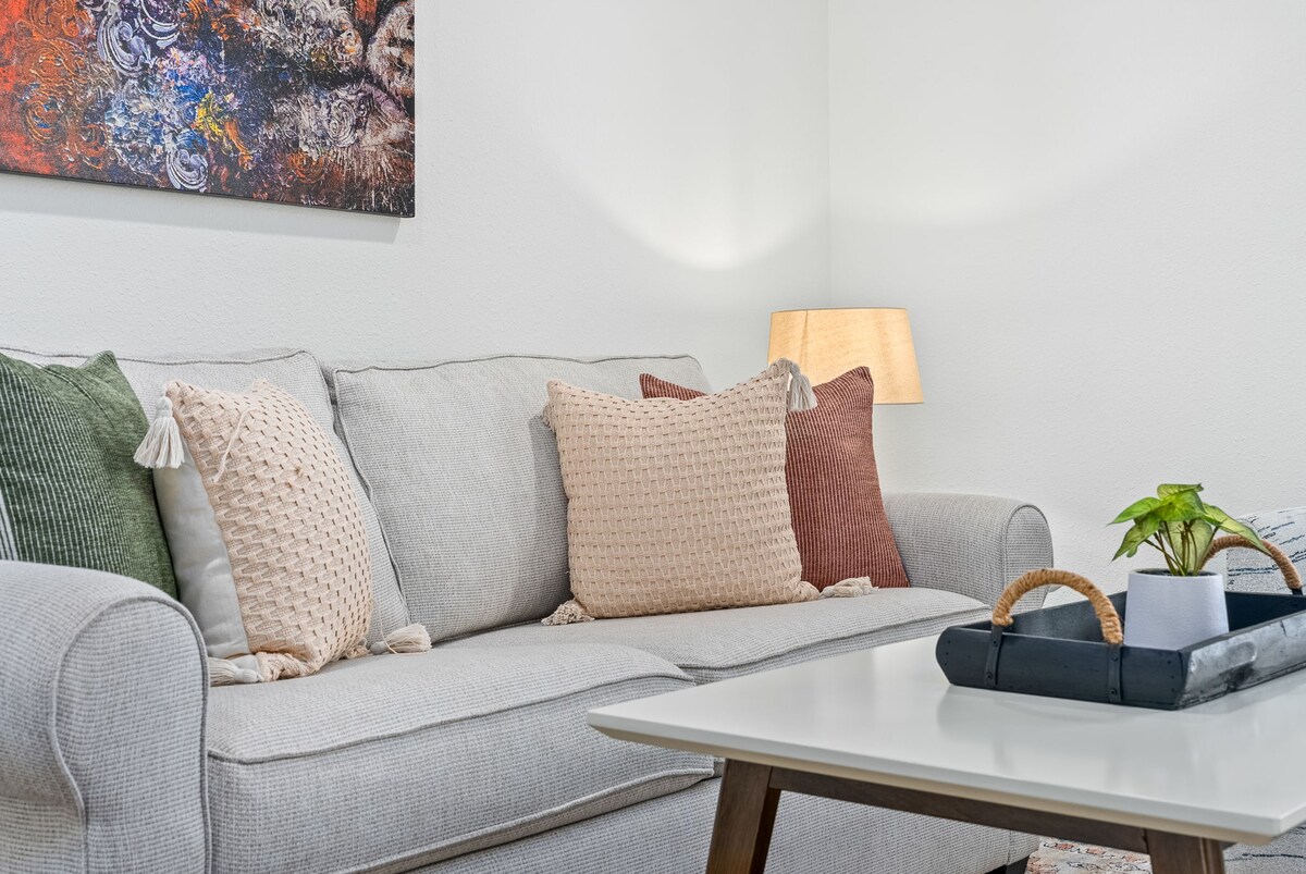 A light-colored sofa is adorned with decorative pillows in soft hues. A neutral coffee table with a wooden base is positioned in front, featuring a small potted plant. A warm lamp provides soft lighting, and abstract artwork is displayed on the wall.