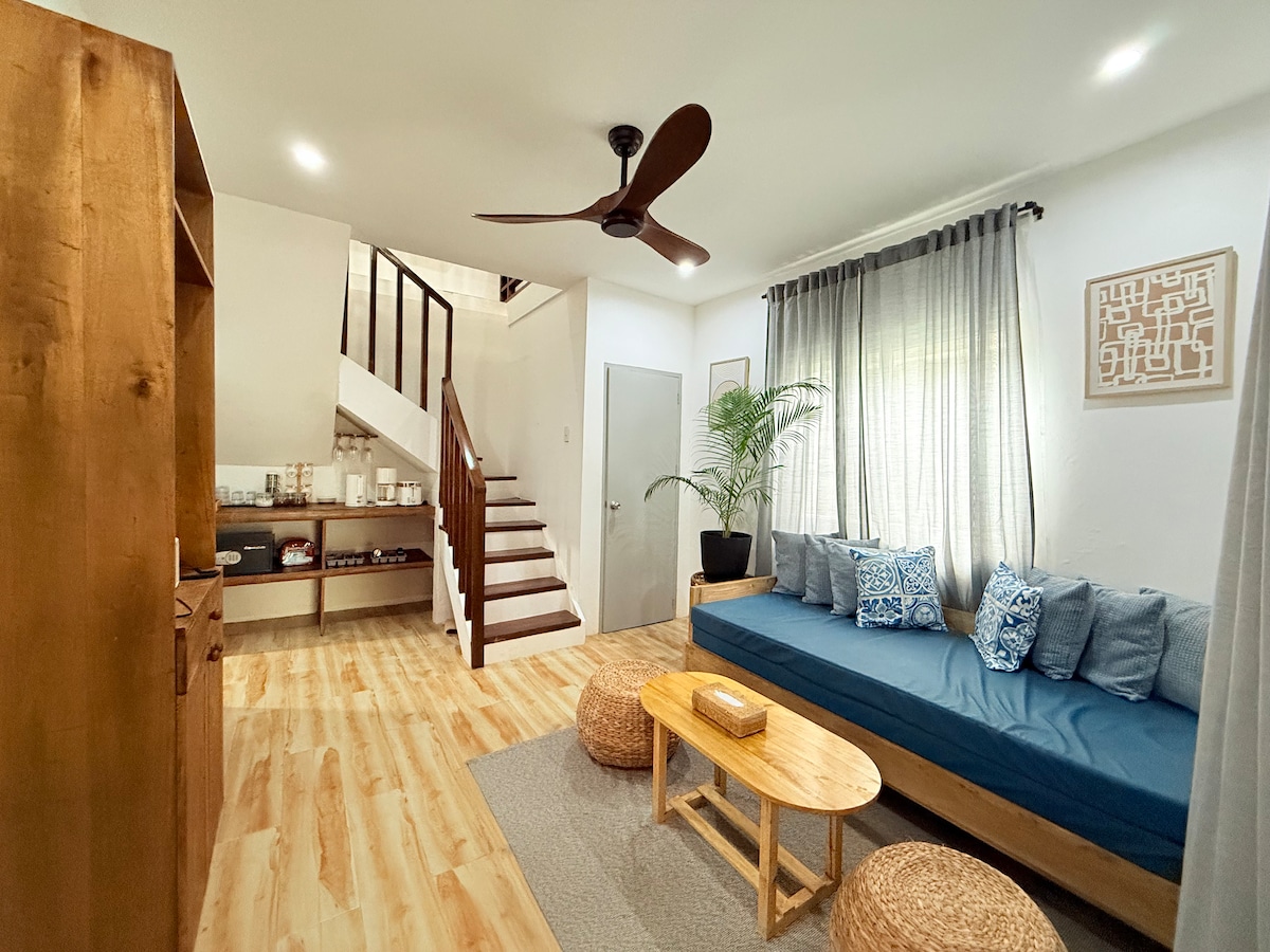 A spacious living area is arranged with a cozy, blue sofa adorned with decorative pillows, accompanied by two wooden coffee tables. The room features light wood flooring and is illuminated by natural light, with a staircase leading to the upper level and potted greenery adding a touch of nature.