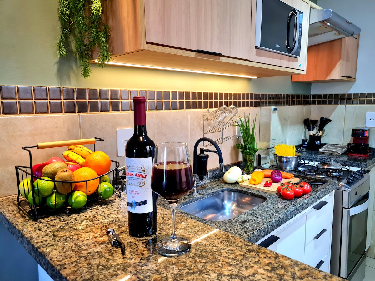 A modern kitchen features a granite countertop with a variety of fresh fruits displayed in a wire basket. A bottle of wine and a glass sit beside an assortment of colorful vegetables, pasta, and an assortment of cooking utensils. Appliances include a microwave and stovetop.