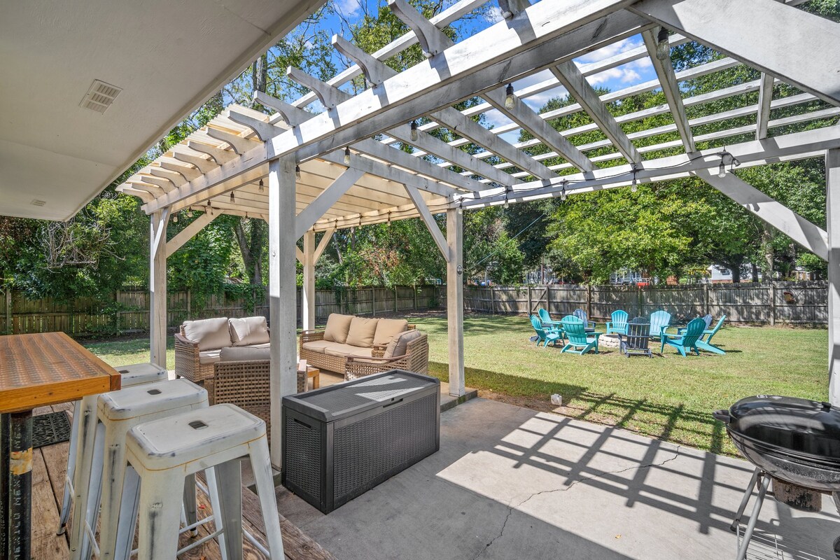An inviting outdoor space features shaded seating beneath a pergola, complemented by a comfortable sofa and a modern fire pit. Colorful Adirondack chairs are positioned across the grassy area, providing a relaxed setting for gatherings. Well-maintained greenery surrounds the space, enhancing the natural feel.