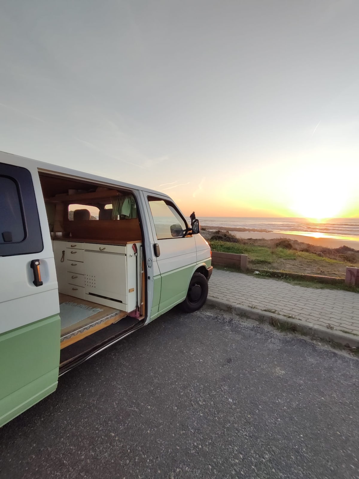 The side of a VW T4 campervan is shown, with its doors open towards a beach at sunset. The soft colors of the sky reflect on the water, while lush greenery and a sandy shore are visible in the background.