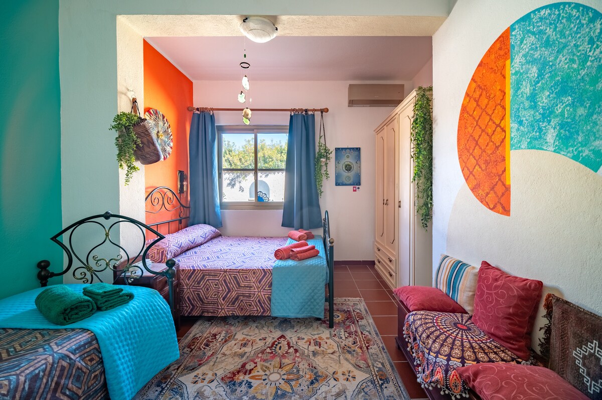 A bright and spacious triple bedroom features decorative walls in vibrant shades of orange and teal. Two beds are neatly arranged, adorned with colorful textiles. A large window allows natural light to fill the room, complemented by decorative plants and a cozy seating area with patterned cushions.