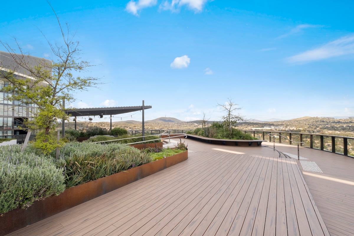 An expansive outdoor terrace features smooth wooden decking and a variety of greenery. A sheltered seating area is visible, along with open views of the surrounding landscape and sky, creating a serene space for relaxation.