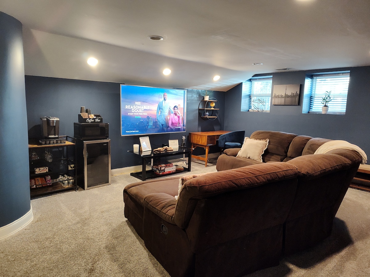 A cozy entertainment area is shown with a large screen displaying a movie. Comfortable seating is arranged in a semi-circle, with a mini fridge and coffee station nearby. Soft lighting enhances the atmosphere, and light filters in through small windows with blinds.