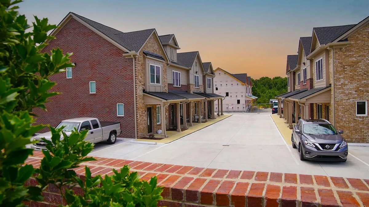 A view of the Pinewood Place townhomes is presented, showcasing two rows of well-appointed brick buildings. The paved driveway accommodates parked vehicles, while lush greenery lines the entrance, with a vibrant sunset casting a soft glow over the scene.