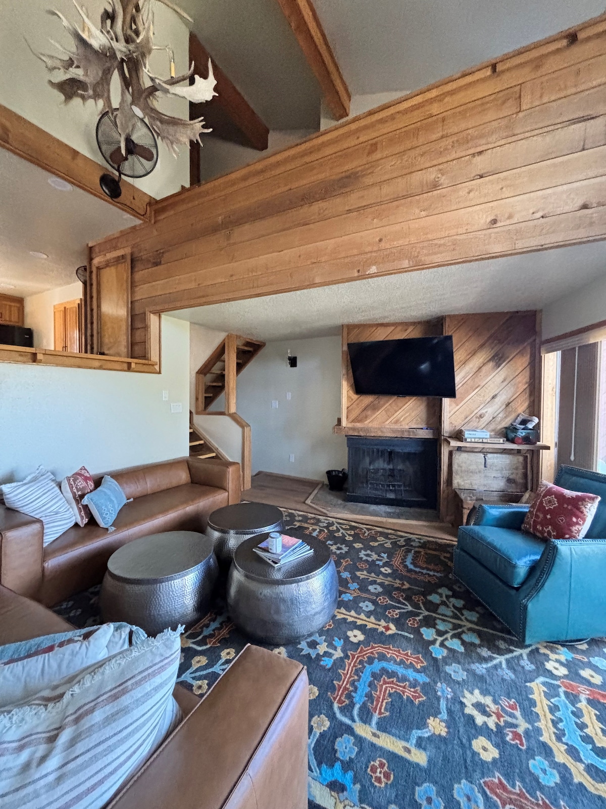 A warm living area features high wooden beams and a cozy seating arrangement. A large, decorative rug covers the floor, complementing two metallic tables. A fireplace stands against the wall beneath a flat-screen TV, with stairs leading to an upper level in the background.