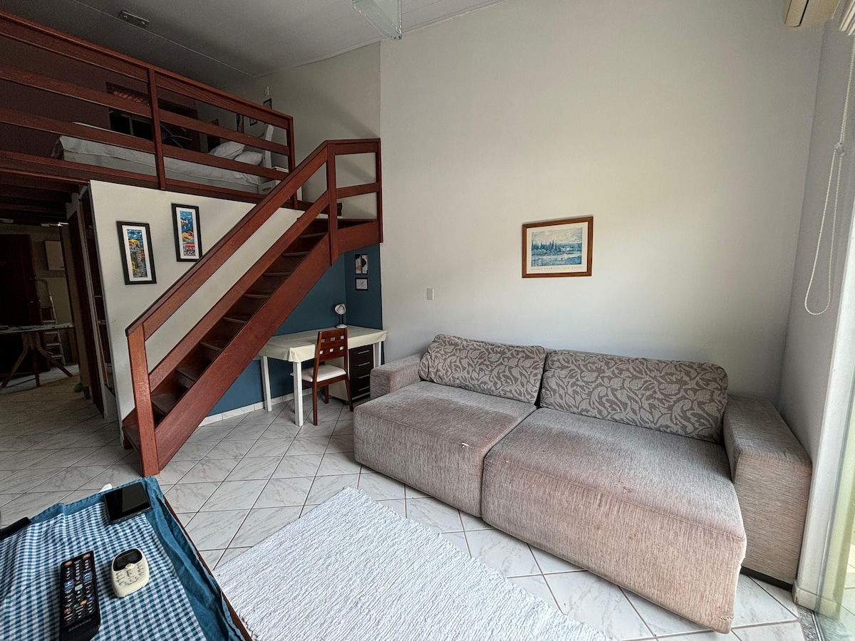 A spacious living area features a comfortable sofa and a small dining table. A decorative artwork hangs on the wall. Stairs lead to a lofted sleeping area, accessible from the living space. Natural light enters through large windows, enhancing the open atmosphere.