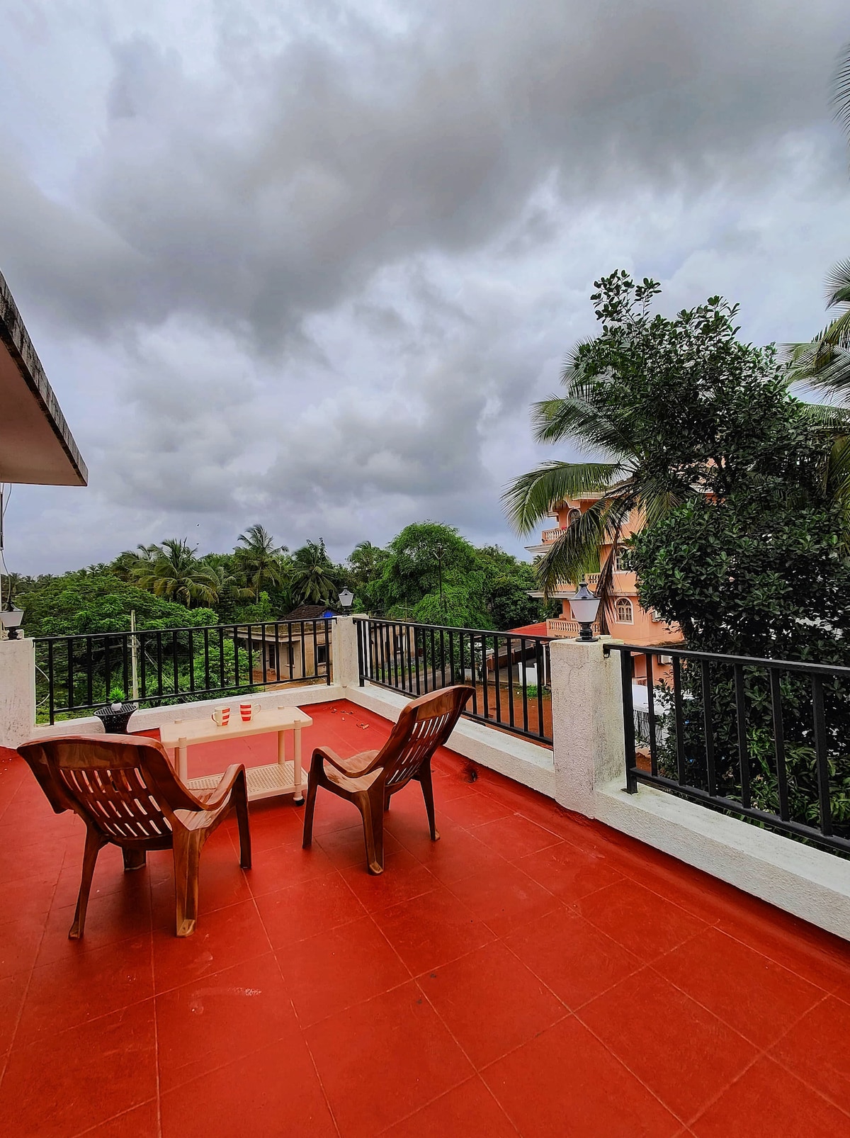 A private terrace features two brown chairs and a small table. The red tiled floor contrasts with the greenery surrounding the area. Dark clouds float above, creating a serene atmosphere amidst the trees and buildings in view.