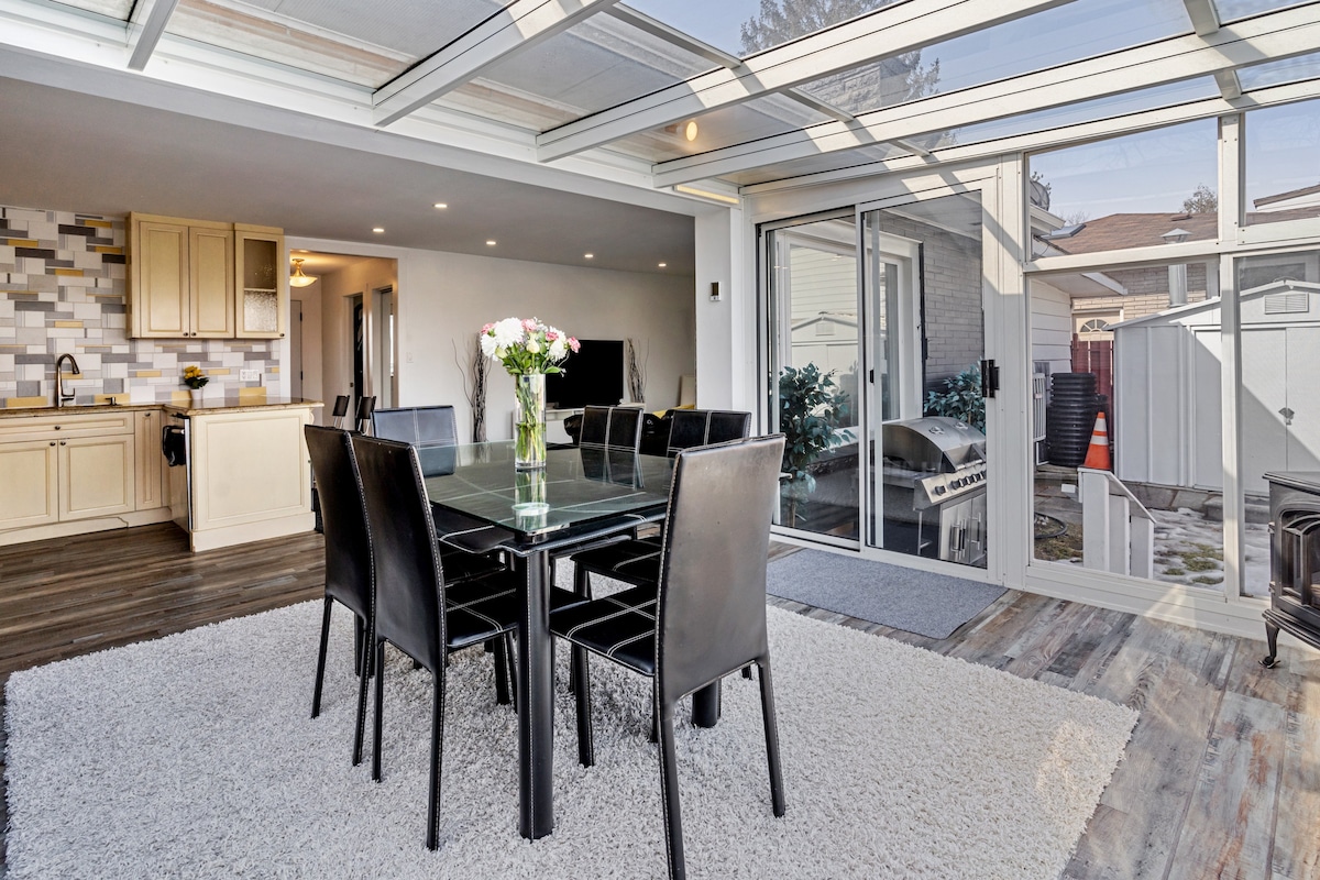 A dining area is featured with a glass-top table surrounded by black chairs. Natural light floods the space through sliding glass doors that lead outside, while a soft area rug adds comfort underfoot. The kitchen area is partially visible in the background.