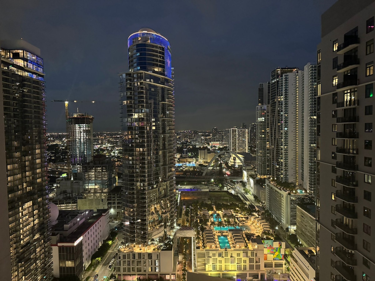 The nighttime cityscape showcases a vibrant skyline, with tall buildings illuminated in various colors. The lush greenery and a large swimming pool can be seen below, adding a tranquil touch to the bustling urban environment.