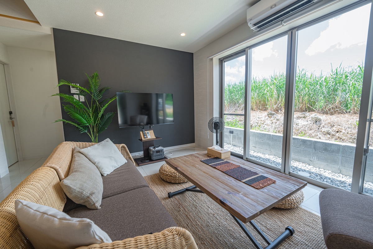 A seating area features a comfortable sofa adorned with various cushions, complemented by a wooden coffee table. A television is mounted on the wall, and large sliding glass doors provide a view of the lush sugarcane fields outside, allowing natural light to fill the space.